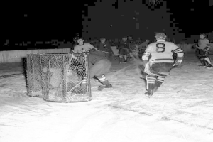 Schwarzes und weißes Bild von Männern, die Hockey auf einem Eisplatz mit einem Netz im Vordergrund und einer Wand im Hintergrund spielen.