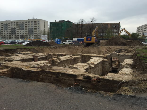 Baustelle mit einem großen Erdwall, Baggern, Fahrzeugen, Gras, Bäumen, Gebäuden mit Fenstern, bewölktem Himmel und einer Straße im Vordergrund, wahrscheinlich in Berlin.