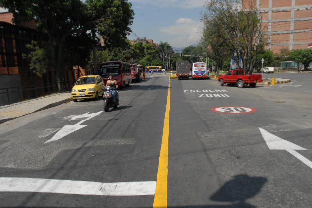 Eine Straßenszene mit mehreren Gebäuden, Bäumen, fahrenden Fahrzeugen, Schildern, Verkehrsampeln an Pfählen, einem blauen bewölkten Himmel und Warnschildern auf der Straße.