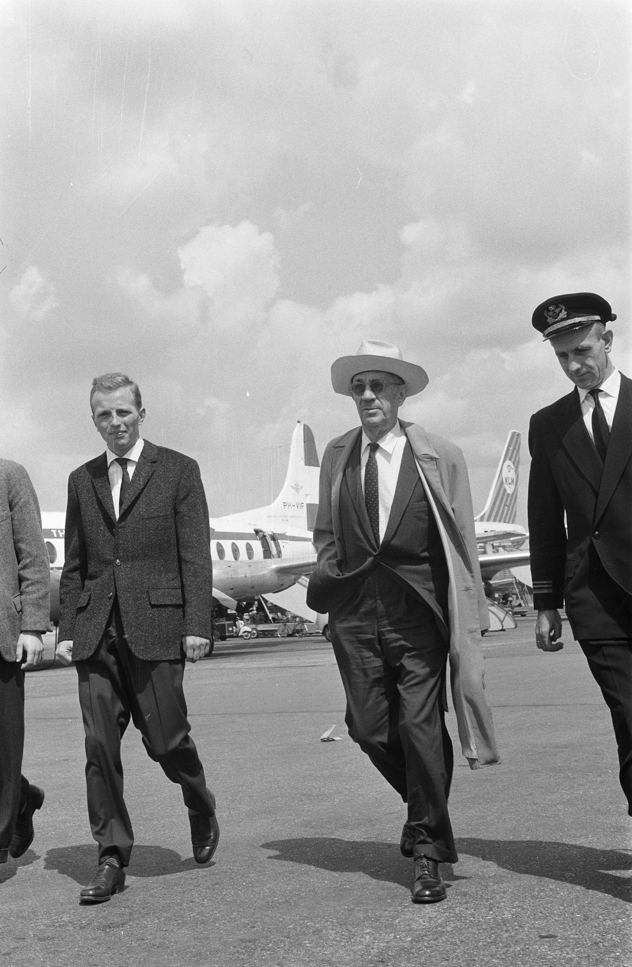 Eine Gruppe von Männern in Anzügen und Hüten geht auf ein Flugzeug auf dem Rollfeld zu, mit einem klaren Himmel im Hintergrund, dargestellt in Schwarz-Weiß.