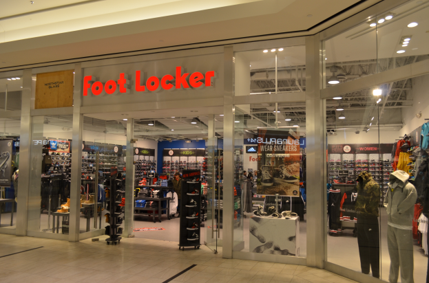 Ein Foot Locker-Laden in einem Einkaufszentrum mit Glasw√§nden und -t√ľren, Schaufensterpuppen in Kleidern, Regalen mit Schuhen und anderen Artikeln sowie Deckenlampen.