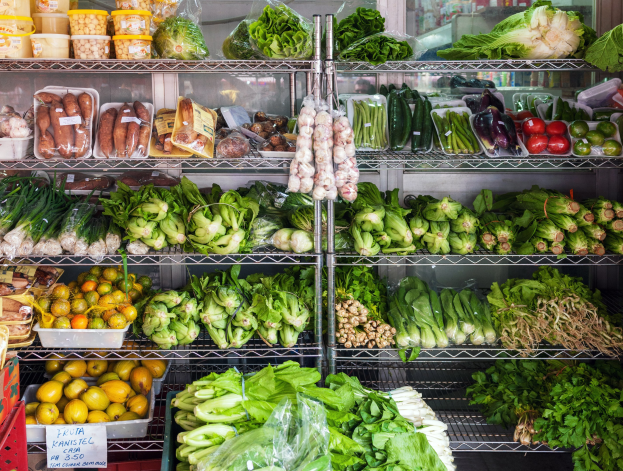 Ein Gang in einem Supermarkt mit frischem Gemüse und Obst in Plastikverpackungen, Kisten und einer Schautafel sowie einem Glasfenster im Hintergrund.