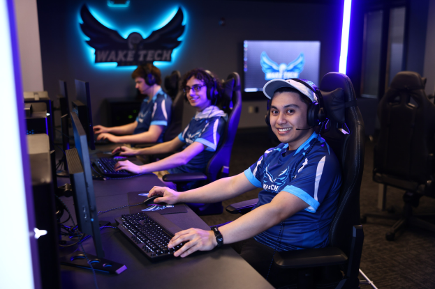 Gruppe von Menschen an einem Schreibtisch mit Computern, Kopfhörern und strahlenden Gesichtern, mit Monitoren, Tastaturen und anderen Gegenständen auf dem Schreibtisch, einem Logo und Text an der Wand dahinter und Stühlen rechts.