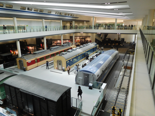 Ein großer Museumsraum mit verschiedenen Zügen in unterschiedlichen Größen und Farben, mit Menschen, die die Ausstellungsstücke beobachten und Deckenbeleuchtung.