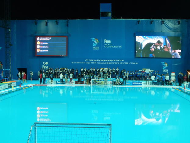 Gruppe von Menschen um ein Hallenbad mit Netz am Boden herumstehend, mit einer Wand im Hintergrund, die "FINA World Championships 2015 Kazan"-Text und Logos anzeigt, sowie Tr├Ągern und Lichtern.