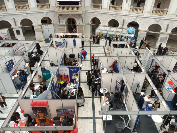 Eine große Gruppe von Menschen versammelt sich in einem Raum mit Ständen, Bannern, Tischen, Stühlen und Luftballons, mit einem Gebäude im Hintergrund, das Bögen, Säulen, Geländer und einen Bildschirm aufweist.