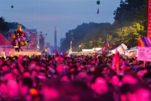 Eine große Menge bei einem nächtlichen Musikfestival mit farbigen Lichtern, Ballons, Zelten, Bannern, Fahnen, Bäumen, einem Turm und einem sternenklaren Himmel im Hintergrund.