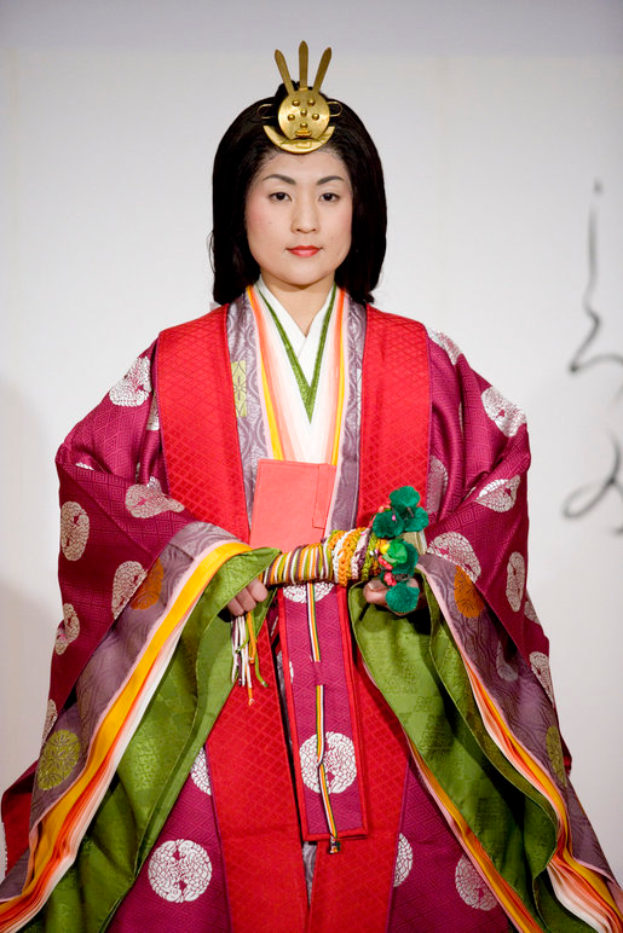 Eine Frau in einem traditionellen japanischen Kimono, das ein leuchtendes rotes und grünes Kleid mit kunstvollen Mustern trägt, steht selbstbewusst auf einem Laufsteg, ihr Haar ist ordentlich zu einem Knoten gebunden.
