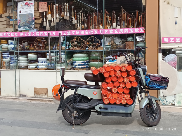 Ein Motorroller mit einem Korb ist auf der Straße vor einem Laden mit einer Glaswand geparkt, auf der verschiedene Gegenstände wie Schalen und Teller ausgestellt sind, mit einer Sichttafel im Inneren.