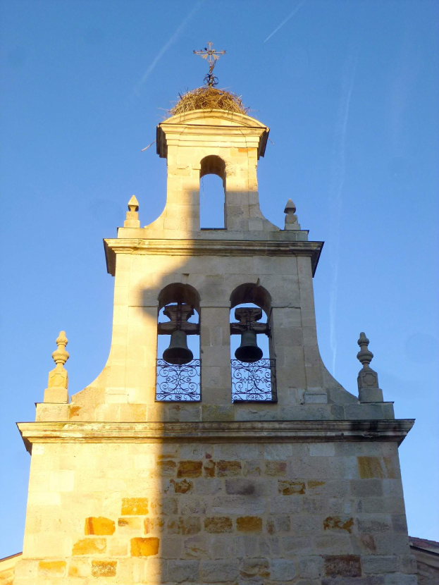 Ein Kirchturm mit einem Kreuz darauf, umgeben von einem Gebäude und einem klaren blauen Himmel, mit einer Glocke, die mit Glocken und einem Geländer verziert ist.