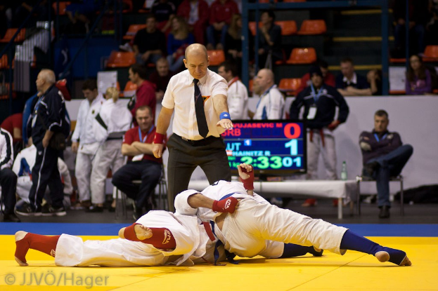 Zwei Judoka im Kampf mit einem Schiedsrichter, der zusieht, während Zuschauer und Offizielle im Hintergrund des Indoor-Veranstaltungsorts zu sehen sind.