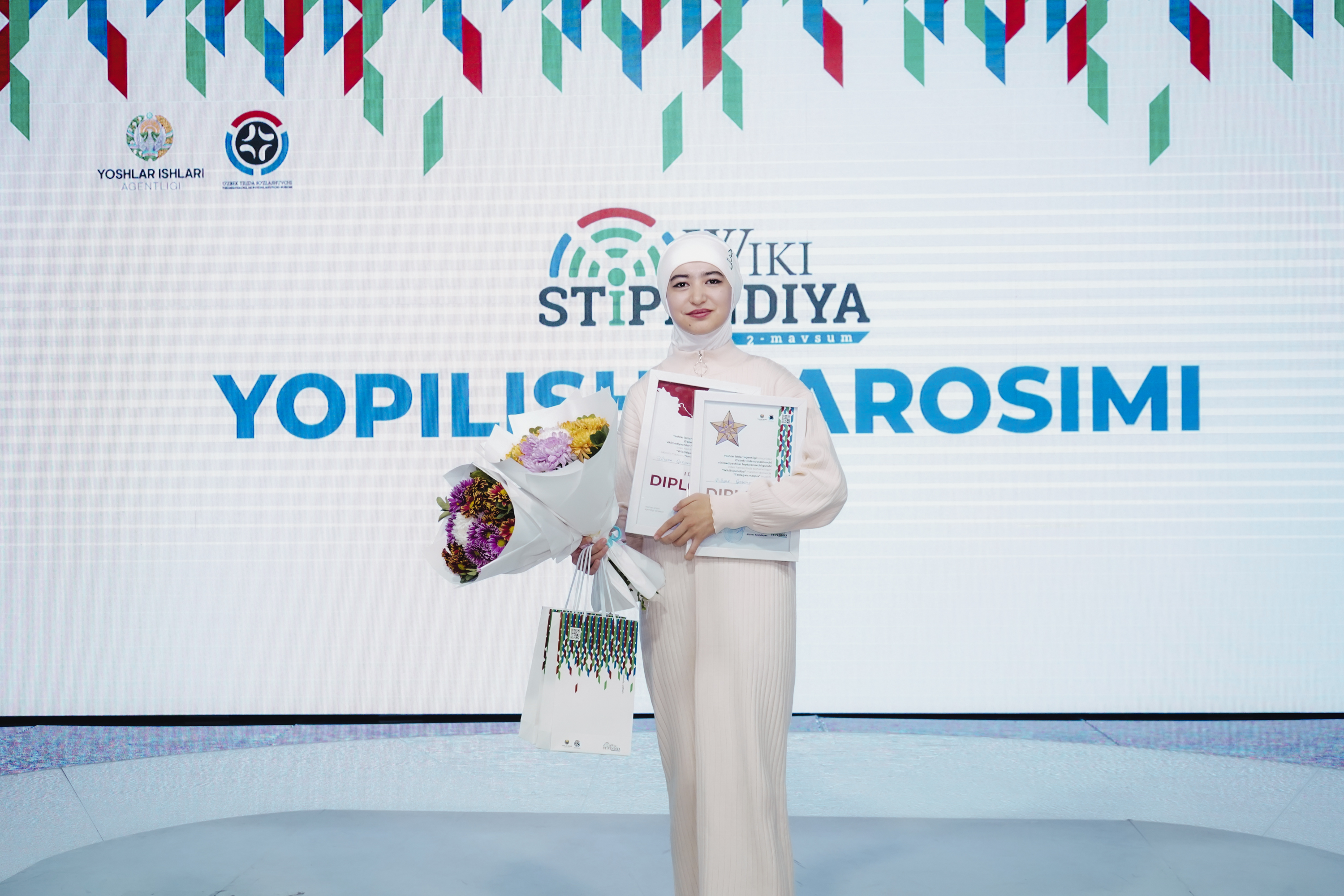 Eine Frau steht auf einer Bühne und hält in einer Hand einen Blumenstrauß und in der anderen ein Zertifikat, während hinter ihr ein Banner mit der Aufschrift "Yopilisi Sarosimi" hängt, was darauf hinweist, dass sie die Gewinnerin des Yopilis Sarosimi Awards ist.