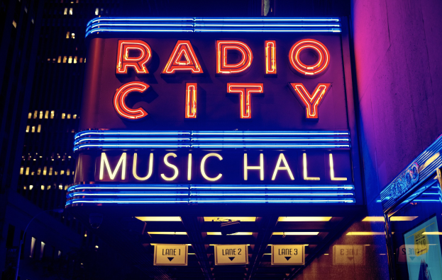 Das beleuchtete Radio City Music Hall in New York City, umgeben von hohen Gebäuden, mit einer "Radio City"-Leuchtschrift und zusätzlichen Schildern auf der rechten Seite.