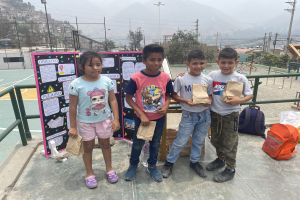 Gruppe von Kindern auf einem Basketballfeld mit Papiertüten, Vorräten auf dem Boden und einer Tafel mit Text links daneben, vor einem Hintergrund aus Zaun, Pfählen, Straßenlaternen, Bäumen, Gebäuden, Hügeln und Himmel.