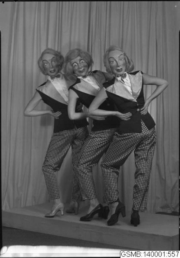 Drei Frauen in Smokings und Fliegen lächeln und posieren für ein Foto auf einer Bühne vor einem Vorhang, das Bild ist in Schwarz-Weiß.