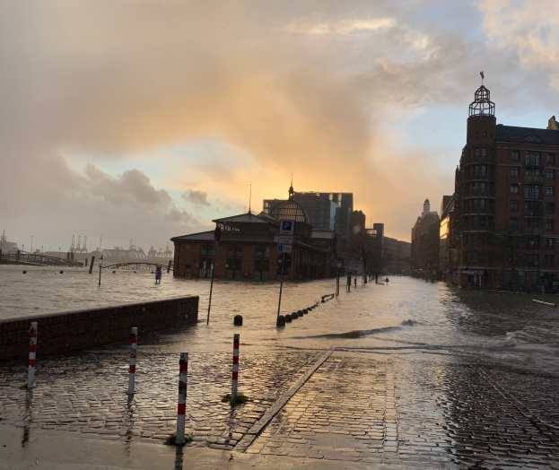 Eine überflutete Straße in Hamburg, Deutschland, mit Wasser auf der Straße, Pfählen, Schildern, Gebäuden mit Fenstern, einer Brücke und einem bewölkten Himmel im Hintergrund.