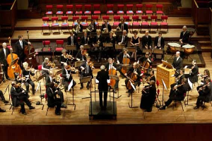 Großes Orchester auf der Bühne mit dirigierendem Musiker, der verschiedene Instrumente spielt, Stühle und Hintergrundtreppe.