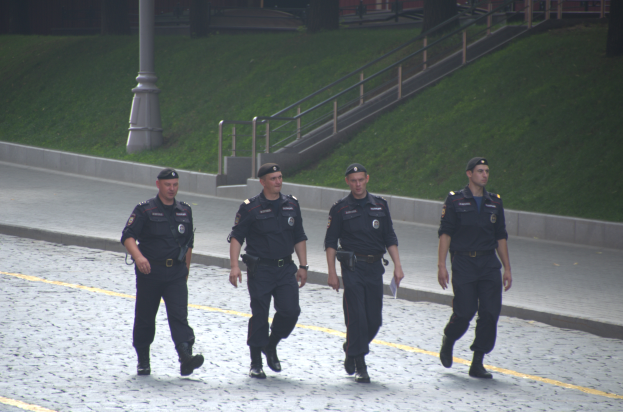 Vier Polizisten in Uniform gehen auf einer Straße und halten Papiere, mit einem Gebäude, Bäumen und einem Pfahl im Hintergrund.