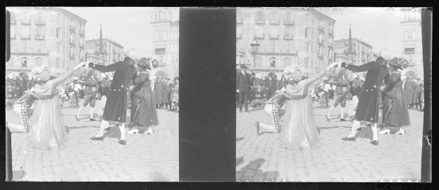 Ein Schwarz-Weiß-Foto einer Gruppe von Menschen in Kostümen, die auf der Straße tanzen, umgeben von Gebäuden, Straßenmasten und Straßenlaternen unter einem klaren Himmel.