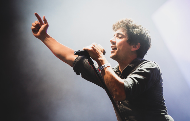 Ein Mann in schwarzem Hemd und Jeans singt in ein Mikrofon auf einer B├╝hne, mit geschlossenen Augen und einem jubelnden Publikum im Hintergrund.