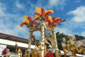 Ein Karnevalswagen, der mit goldenen und roten Federn geschmückt ist, mit Menschen darauf, umgeben von einem Gebäude, Bäumen, Drähten und einem Banner, mit dem Himmel im Hintergrund.