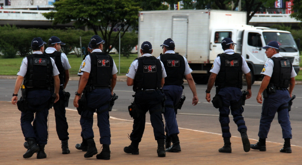 Eine Gruppe von Beamten in Uniform, einschließlich Mützen, Hemden, kugelsicheren Westen, Uhren, Hosen und Schuhen, geht auf einer Straße. Rechts daneben steht ein Lastwagen in der Nähe eines Zauns, mit Gebäuden, Bäumen, Pflanzen, Polen und Bannern im Hintergrund und einer Brücke oben rechts.