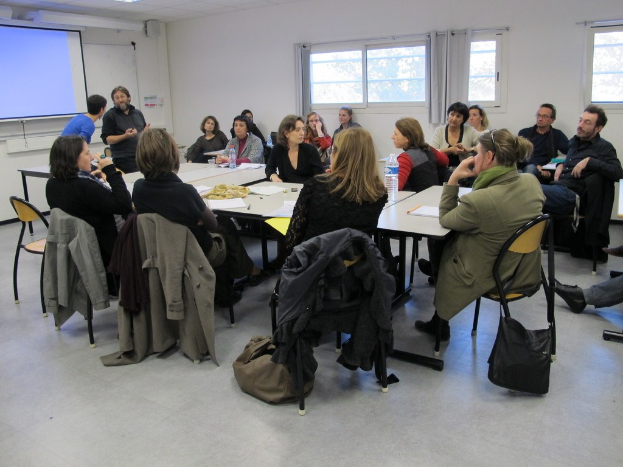 Gruppe sitzender Menschen, die mit zwei stehenden Individuen diskutieren.