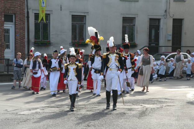 Eine Gruppe von Menschen in Kostümen und mit Stöcken geht eine Straße entlang in einem Umzug, mit Gebäuden, Pflanzen, einer Fahne und Fahrzeugen im Hintergrund.
