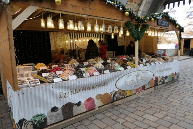 Ein Weihnachtsmarktstand mit verschiedenen Speisen, beleuchtet von Deckenlampen, mit Menschen davor und Bäumen und einem klaren blauen Himmel im Hintergrund.
