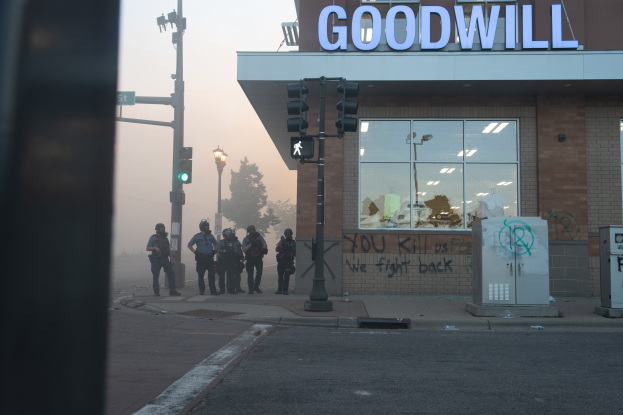 Eine Gruppe von Polizisten in Helmen und mit Waffen steht vor einem Goodwill-Laden mit Glaswänden, mit Laternenmasten, Verkehrszeichen, Bäumen und einem klaren Himmel im Hintergrund.