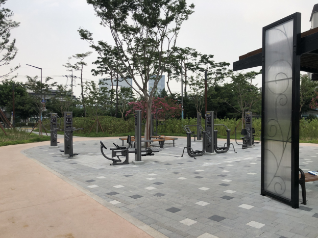 Outdoor-Park mit verschiedenen Fitnessgeräten, Bänken, Bäumen, Pflanzen, Gras, Polen, Lampen, Drähten und Gebäuden vor einem Himmel-Hintergrund.
