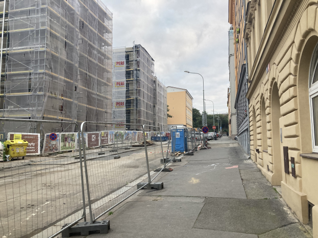 Eine Stadtstraße mit Gebäuden, Fahrzeugen und einer Baustelle mit Gerüsten im Vordergrund unter einem bewölkten Himmel.