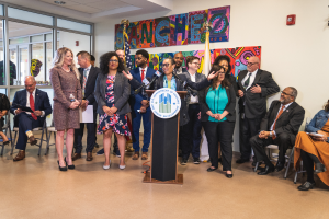 Eine Gruppe von Menschen steht vor einem Podium mit einem Mikrofon und einer Tafel mit der Aufschrift "Sancho Stadtrat tritt dem Stadtrat bei", im Hintergrund sitzen und stehen andere, Fahnen, eine Tafel, ein Feuerlöscher und Deckenleuchten an der Rückwand.