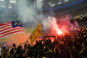 Eine große Menschenmenge in einem Stadion mit Bengalos, Fahnen und einer Kamera, mit Rauch und Schildern mit der Aufschrift "Malaysia vs Malaysia Live-Stream" im Hintergrund.