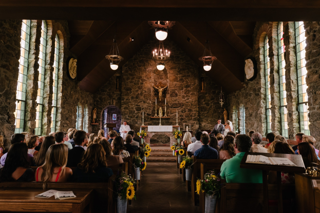 Eine Gruppe von Menschen sitzt in Kirchenbänken während einer Hochzeitszeremonie, mit Blumenschmuck, Büchern und Gegenständen auf den Tischen, Glasfenstern an den Seiten, einer Statue und Kerzen im Hintergrund und Kronleuchtern an der Decke.