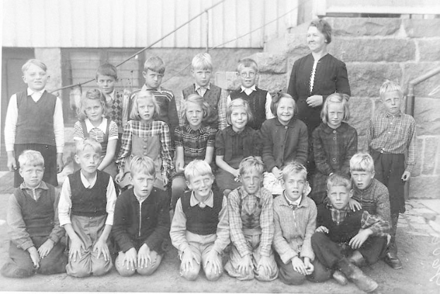 Schwarzes und weißes Foto einer Gruppe von Kindern, die vor einem Gebäude mit Treppe und Geländer posieren, mit der Beschriftung "1930er-Schulkinder" unten.