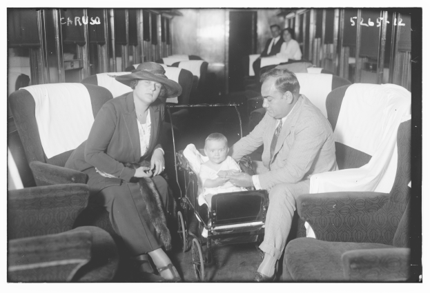 Ein Schwarz-Weiß-Foto eines Mannes im Anzug und einer Frau mit einem Baby, die auf einem Zug mit einem Kinderwagen zwischen ihnen sitzen und andere Passagiere im Hintergrund.