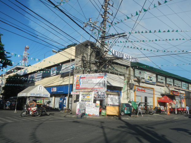 Eine Straßenszene in der Stadt mit einem Telefonmast in der Mitte, umgeben von Gebäuden, Geschäften, Fahrzeugen, Fußgängern, Schirmen, Strommasten, Kabeln, Fahnen, Bäumen und einem bewölkten Himmel.