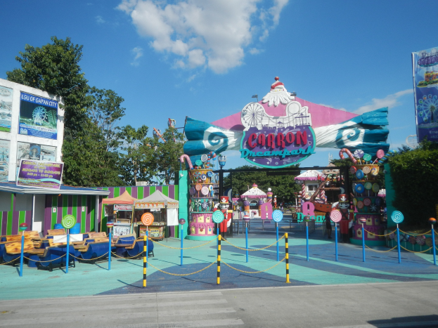 Kirmesfahrgeschäft am Parkeingang mit Straße, eingezäuntem Bereich, Buden, Banner, Bäumen und bewölktem Himmel im Hintergrund.