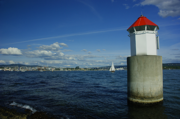 Boote auf dem Meer mit einem Leuchtturm im Vordergrund, Gebäude, ein Berg und der Himmel im Hintergrund.
