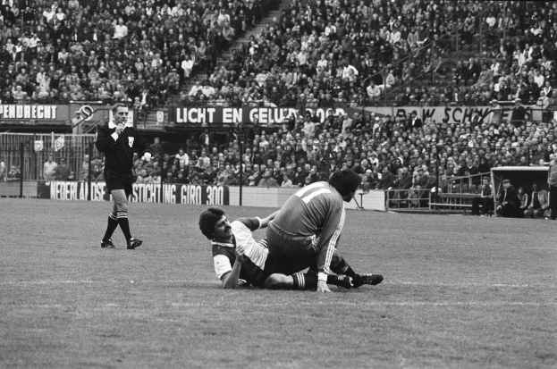 Ein Schwarz-Weiß-Foto von zwei Fußballspielern im Einsatz, wobei einer den anderen angeht, mit Zuschauern und Stadionelementen im Hintergrund und einer Person mit einem Gegenstand auf der linken Seite.
