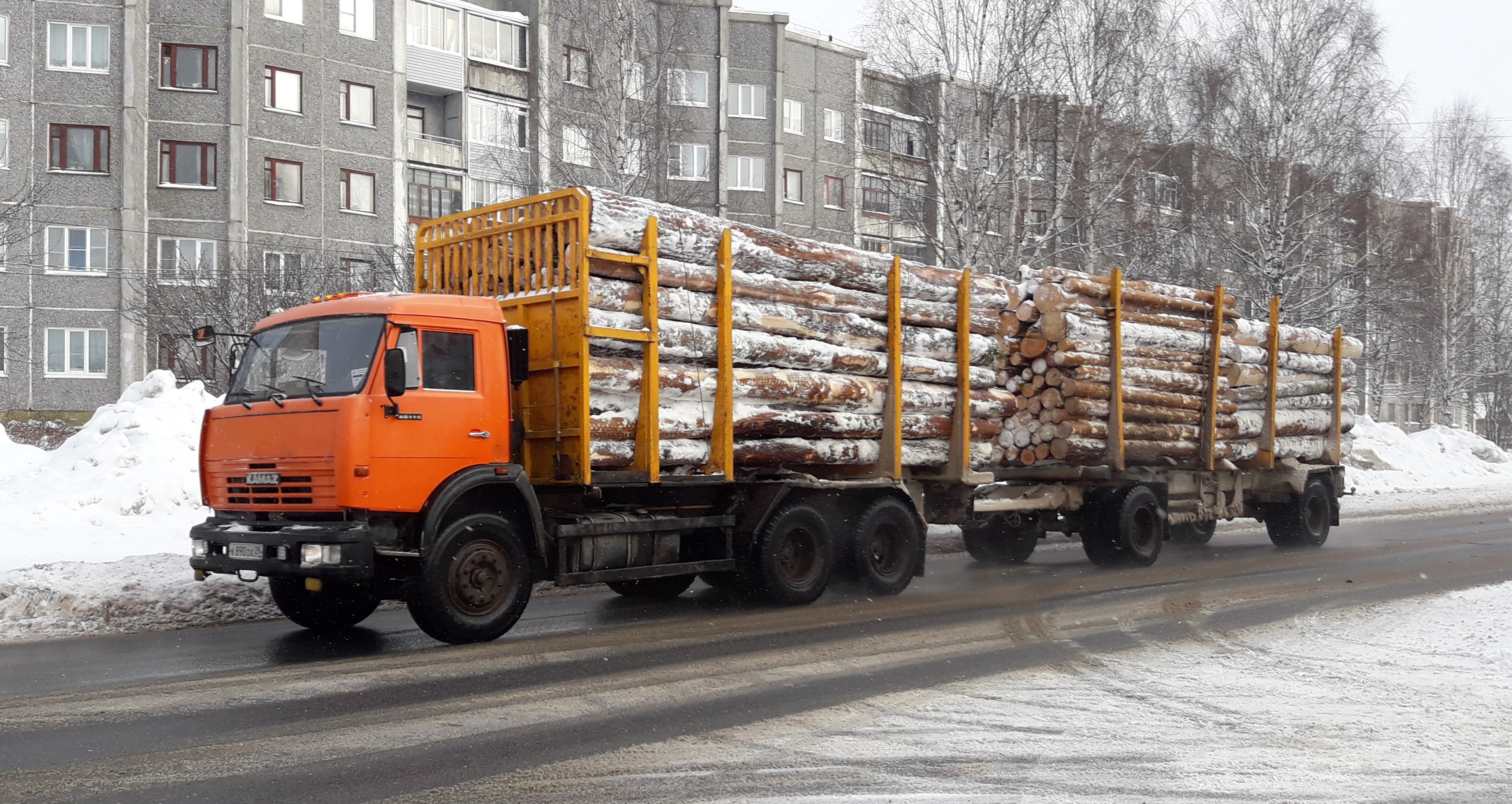 Ein Lastwagen mit Holz fährt auf einer schneebedeckten Straße mit Bäumen und Gebäuden im Hintergrund unter einem klaren Himmel.