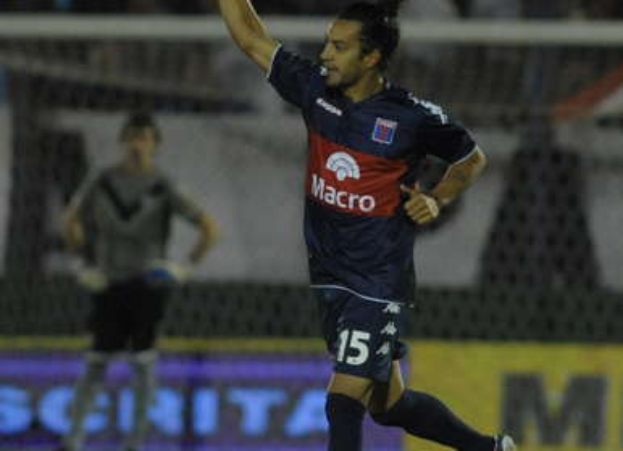 Zwei Fußballspieler feiern ein Tor auf einem Feld, umgeben von einer Gruppe von Menschen, mit einer Person, die eine Kamera hält, einem Banner mit Text, Stühlen und einer Wand im Hintergrund.