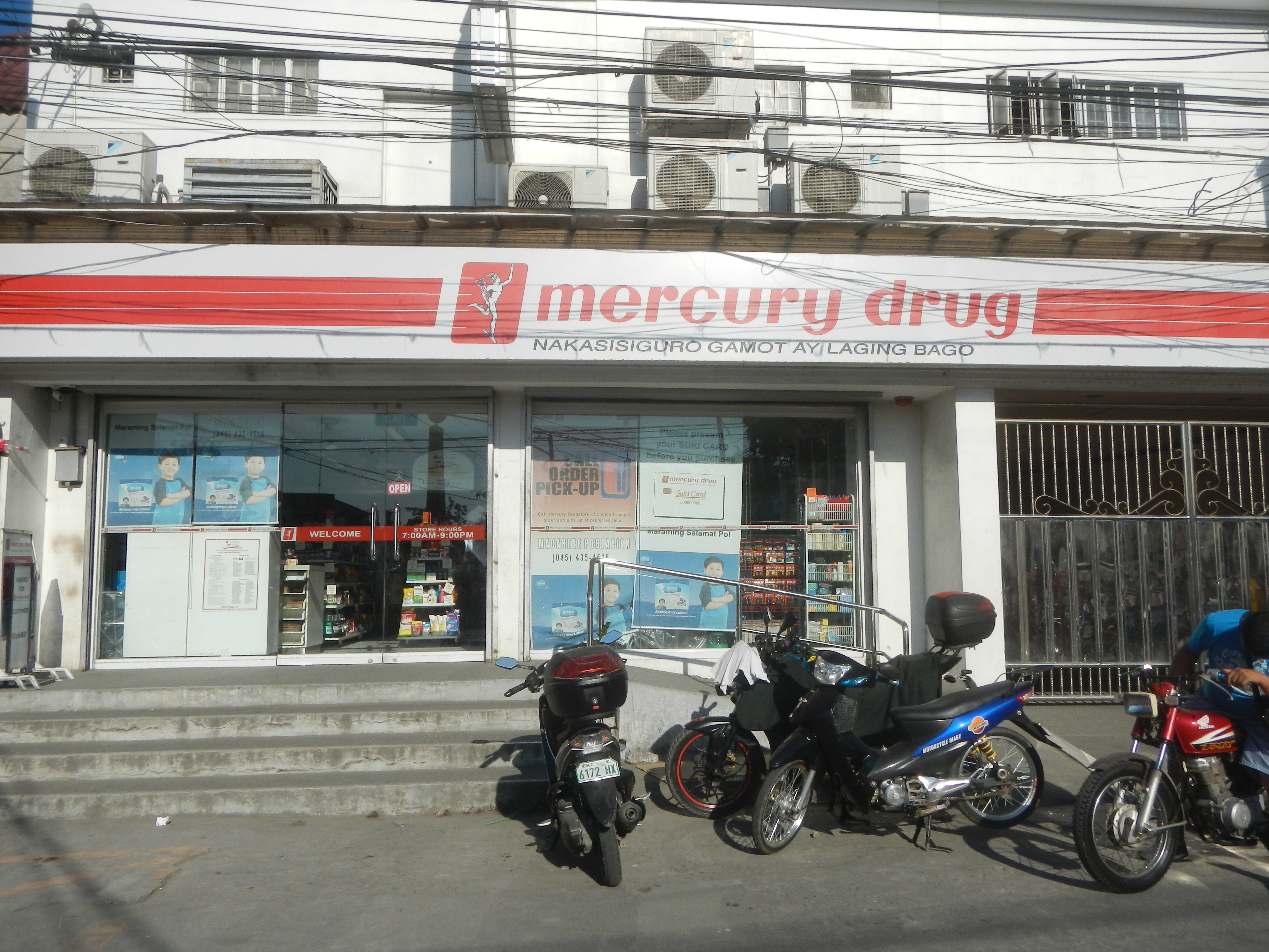Ein Mercury Drug Store mit drei Motorrädern davor, eines mit einer Person darauf, und Gebäuden, Klimaanlagen, Drähten und einem klaren blauen Himmel im Hintergrund.