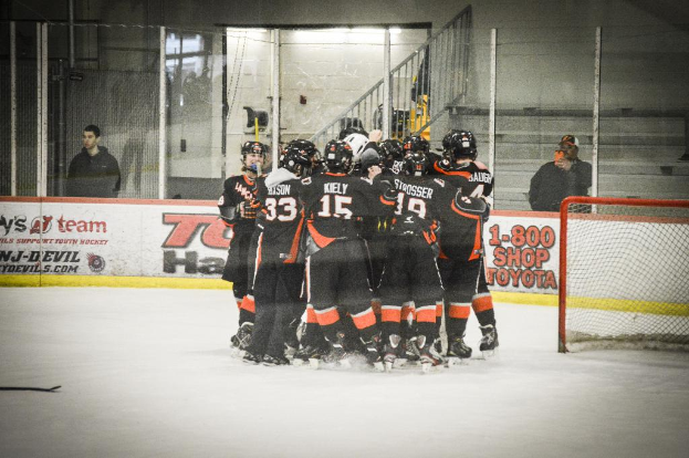 Eine Gruppe von Menschen in Eiskleidung, Handschuhen und Helmen steht auf einem Eisboden mit Absperrungen und Glastüren dahinter, einem Hockey-Tor mit Netz rechts und einem Hockey-Schläger links.