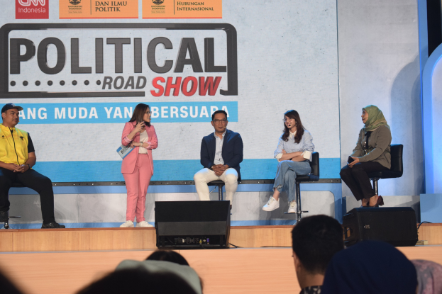 Eine Gruppe von Menschen auf einer Bühne mit einer Frau in der Mitte, die ein Mikrofon hält, eine Zuschauermenge im Vordergrund und ein Banner mit der Aufschrift "Politischer Roadshow 2019" im Hintergrund.