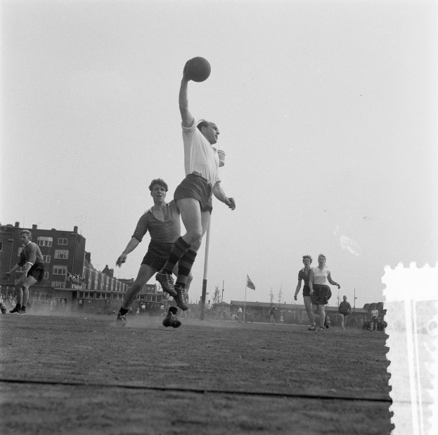 Eine Gruppe von Männern, die Handball auf einem Feld mit Gebäuden, Masten und Fahnen im Hintergrund unter einem klaren Himmel spielen, dargestellt in Schwarz-Weiß.
