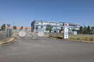 Großes Industriegebäude mit der Aufschrift "First Solar" und vielen Fenstern, umgeben von einem Metallzaun, Bäumen, Pflanzen, Gras, Schildern, Straßenlaternen und einem Weg unter einem bewölkten Himmel.