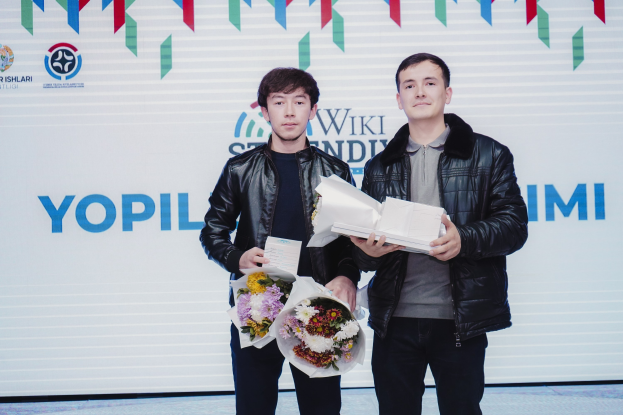 Zwei Männer in schwarzen Jacken stehen auf einer Bühne und halten Blumensträuße, im Hintergrund ein Banner mit der Aufschrift "Yopil International Film Festival", was darauf hinweist, dass sie Festival-Gewinner sind.