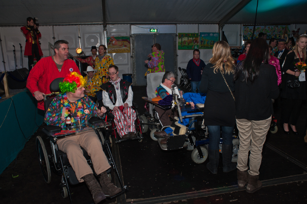Menschen im Rollstuhl in Kostümen, mit einer Menge im Hintergrund, die Gegenstände, Lichter, Rahmen, Bretter, Stangen und ein Tuch hält, unter einem Dach und über einem Boden.